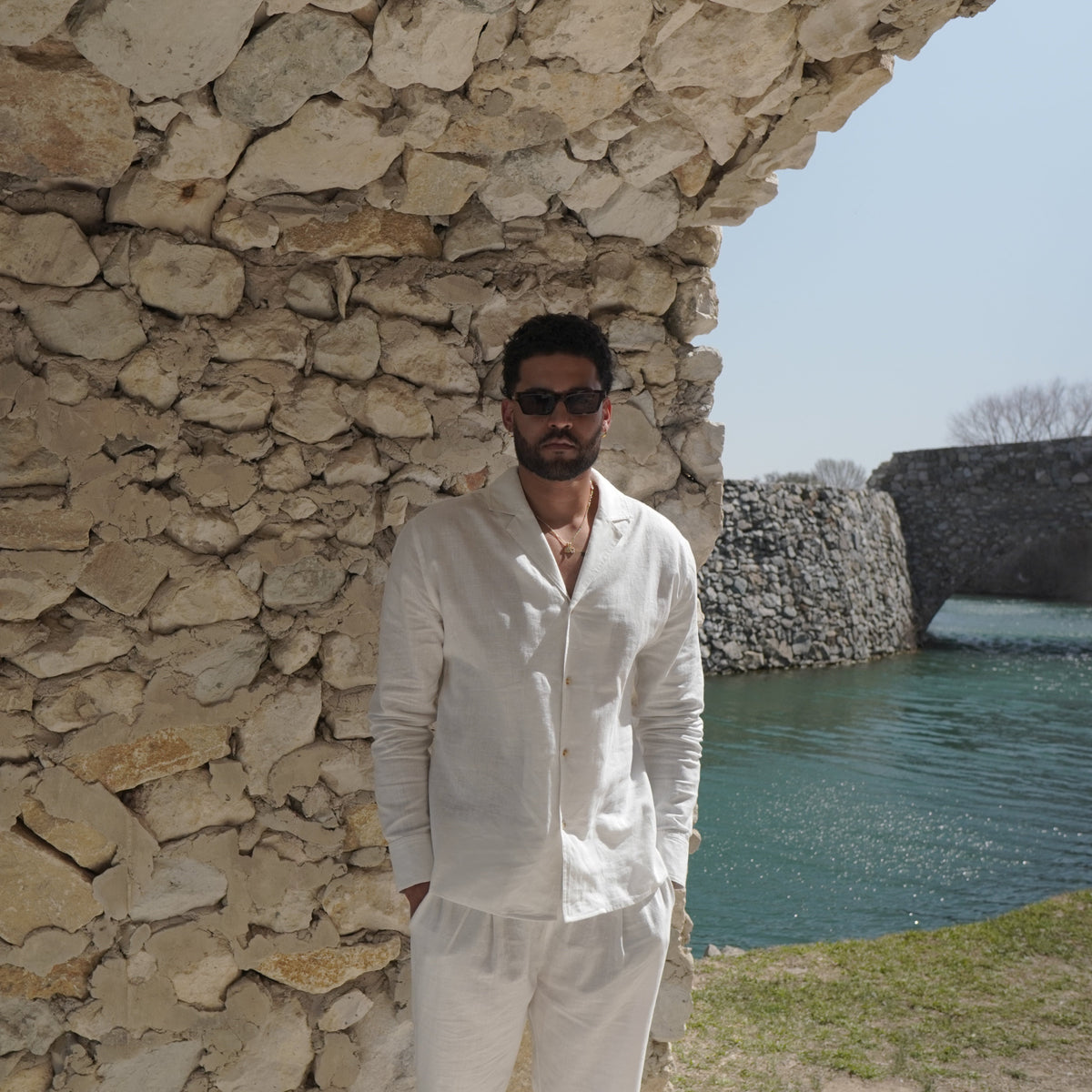 Man in a white outfit standing against a stone wall with water and bridge in the background