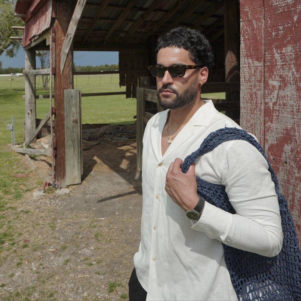 Man in a white shirt and sunglasses leaning out of a wooden structure by a body of water.