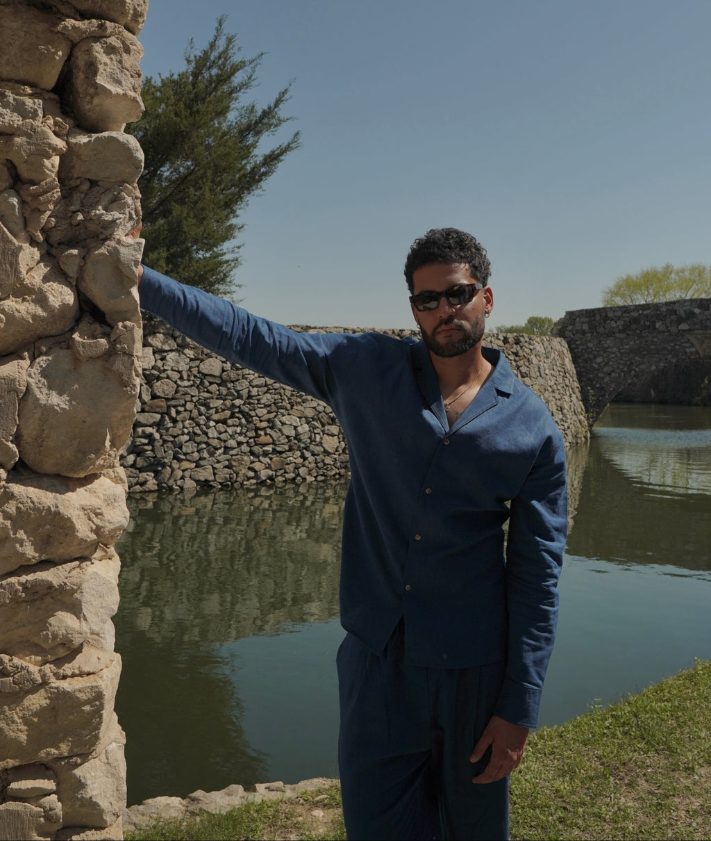 Sher Valor Azure Set – men's slate blue linen shirt and trousers, full body against stone wall with waterfront backdrop
