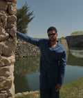 Sher Valor Azure Set – men's slate blue linen shirt and trousers, full body against stone wall with waterfront backdrop