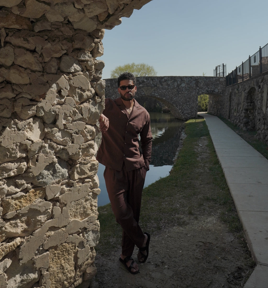 Sher Valor Umber Set – men's dark brown linen shirt and trousers, full body against ancient stone wall with canal backdrop